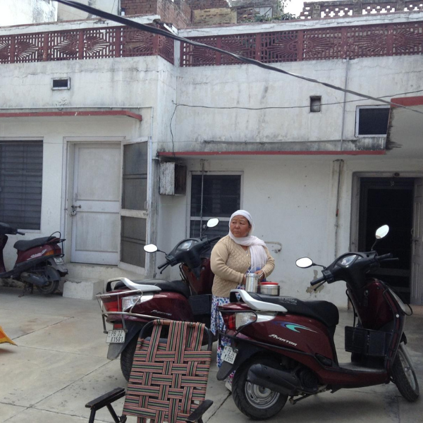 Photo of an Indian hourse, white and with some balconies, and outside a lady standing and a moped